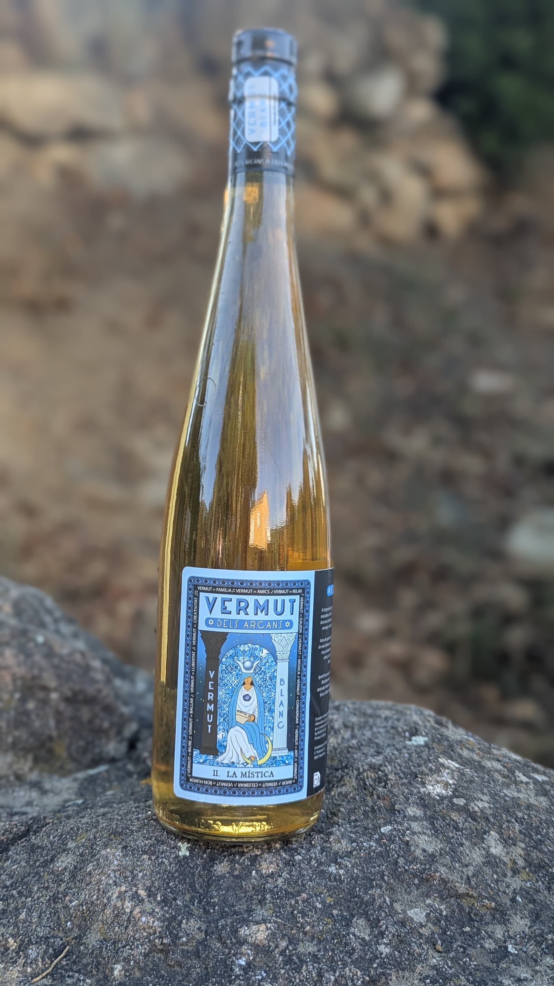 Ampolla La Mística de Vermut dels Arcans sobre roques amb fons de bosc en bokeh
