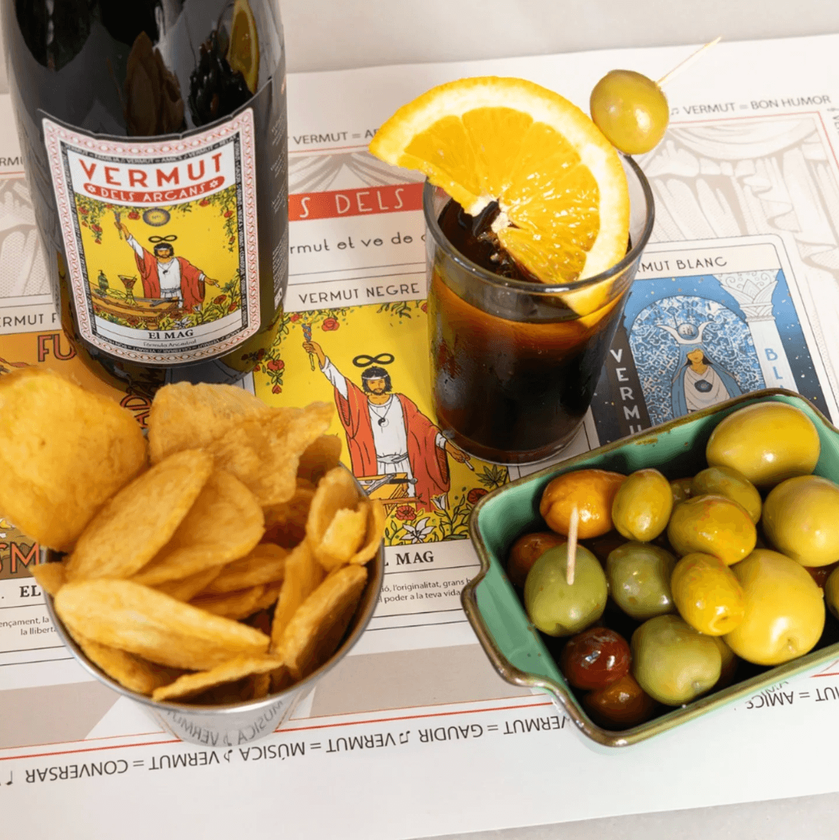 Ampolla El Mag amb copa de vermut, patates i olives sobre l'estovalla dels Arcans