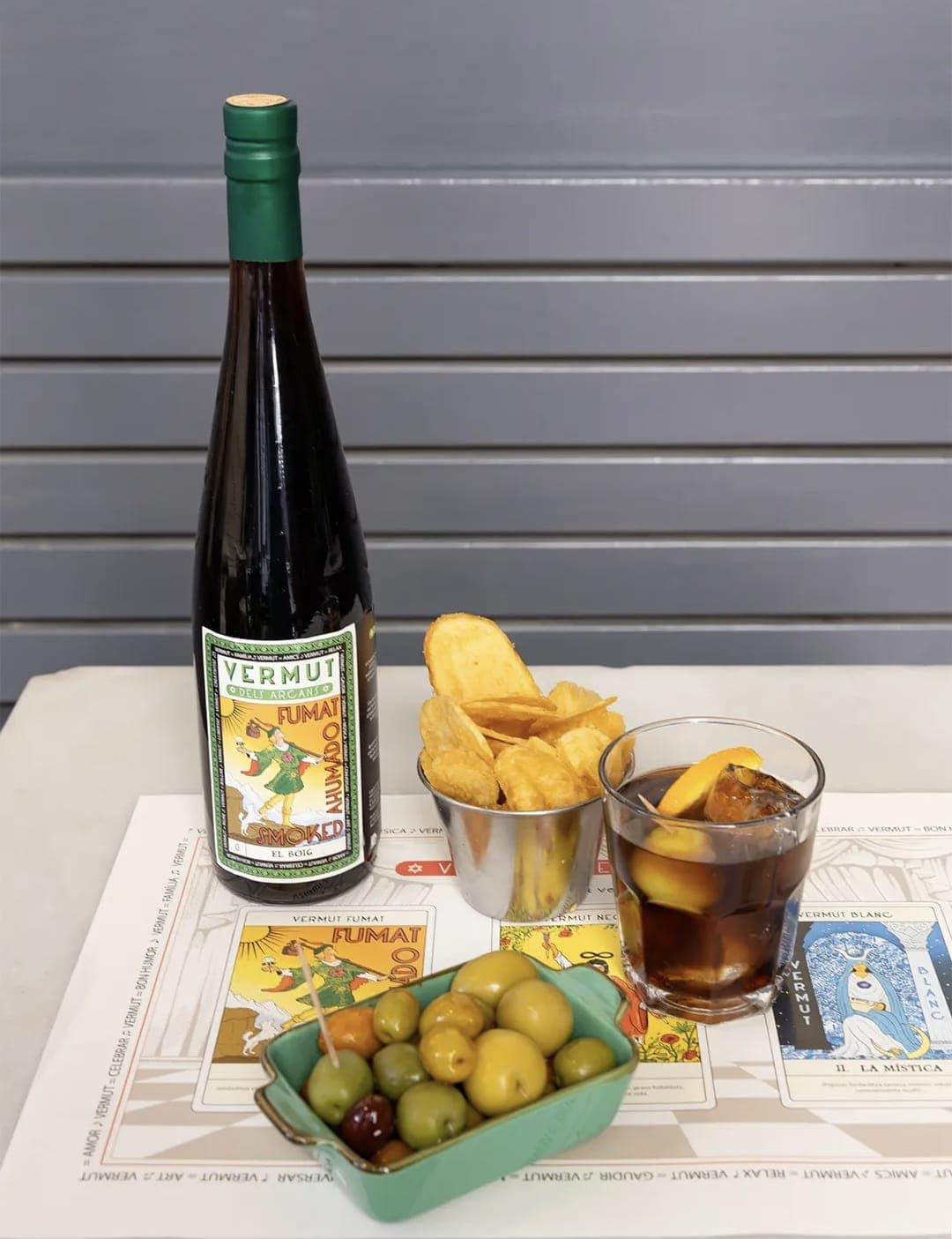 Ampolla Vermut Fumat El Boig amb copa de vermut, patates i olives sobre l'estovalla dels Arcans