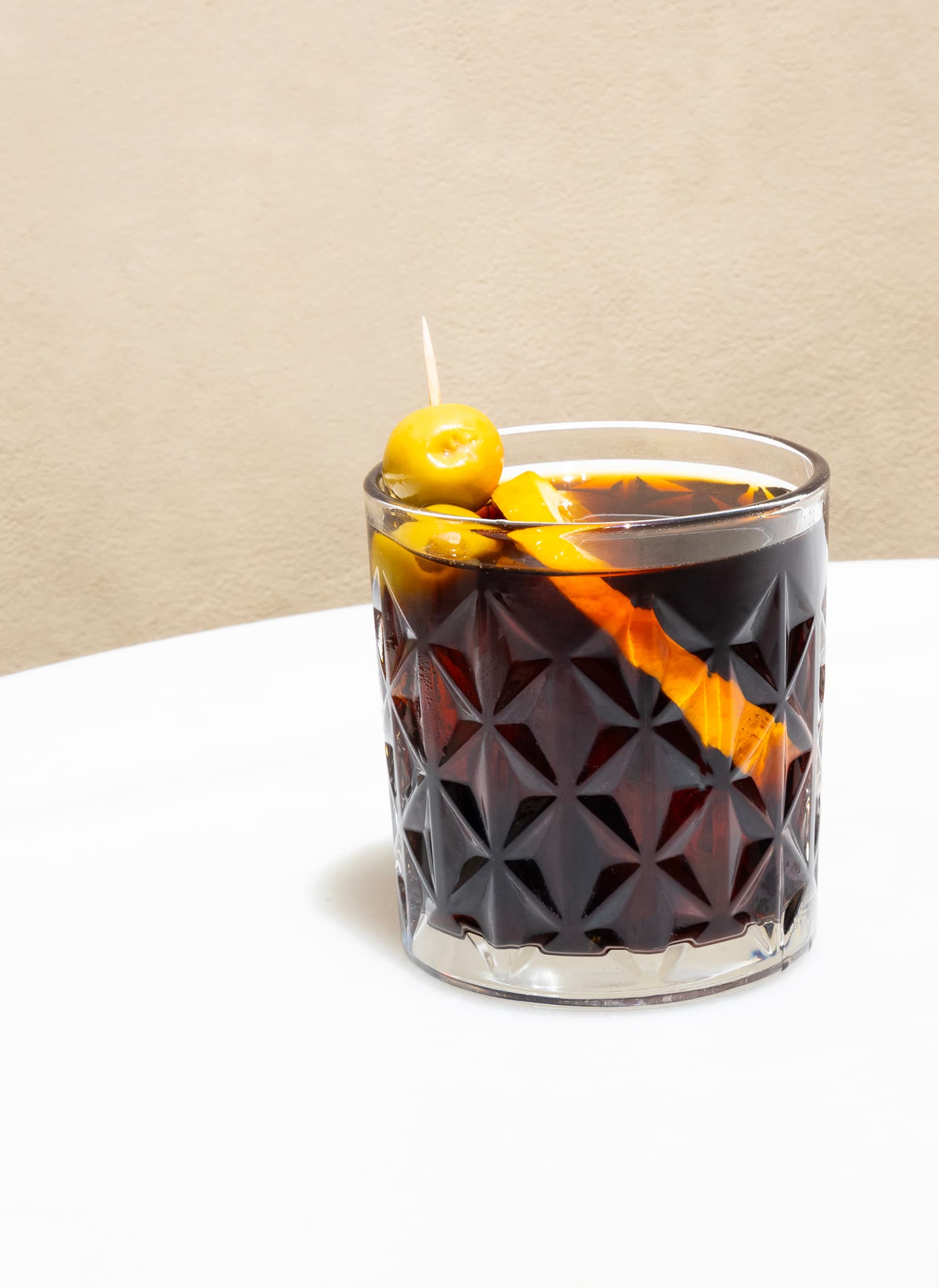 Copa de vermut negre amb pell de taronja i oliva sobre fons beix