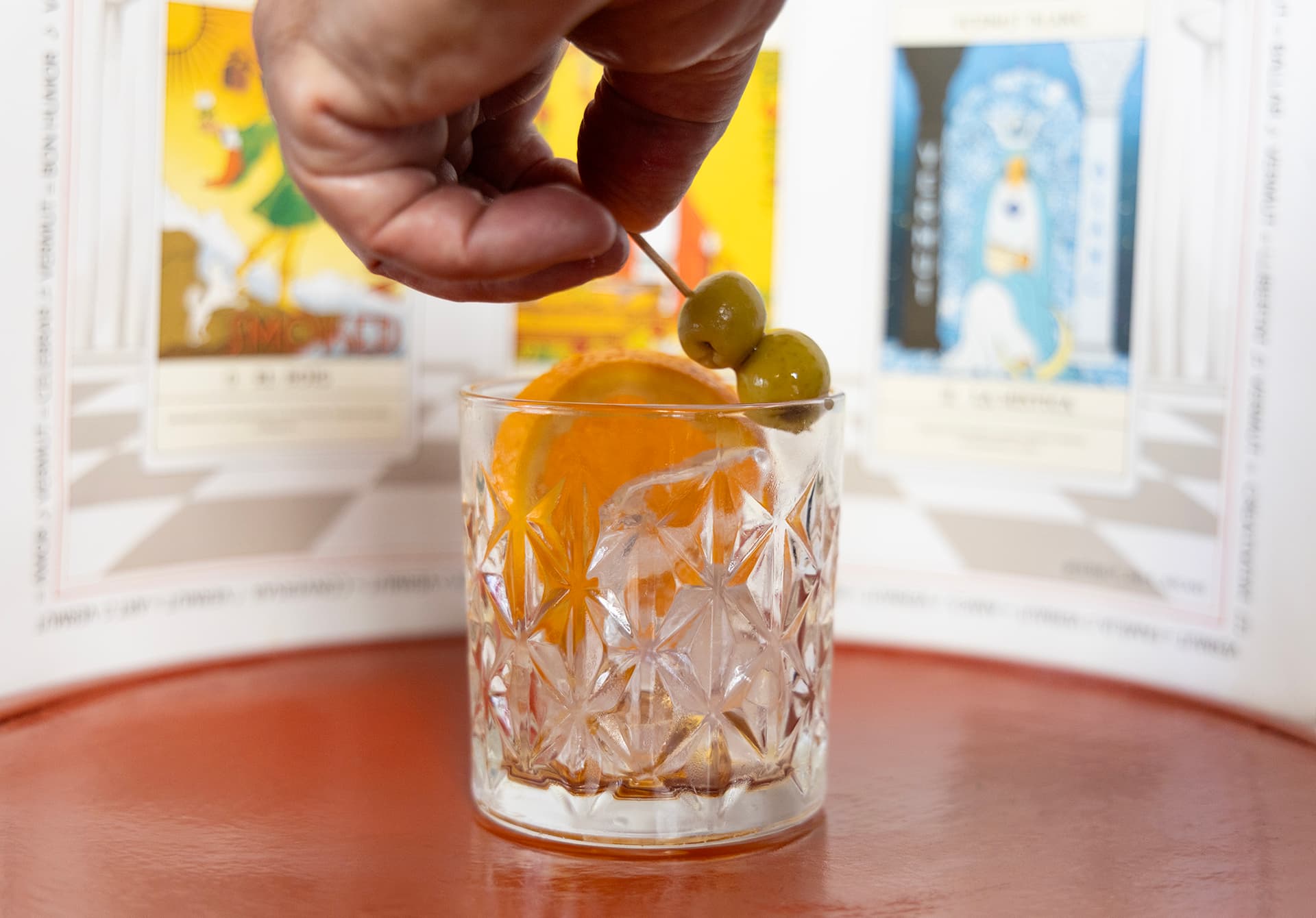 Ma introduint oliva en una copa de vermut blanc amb cartes al fons